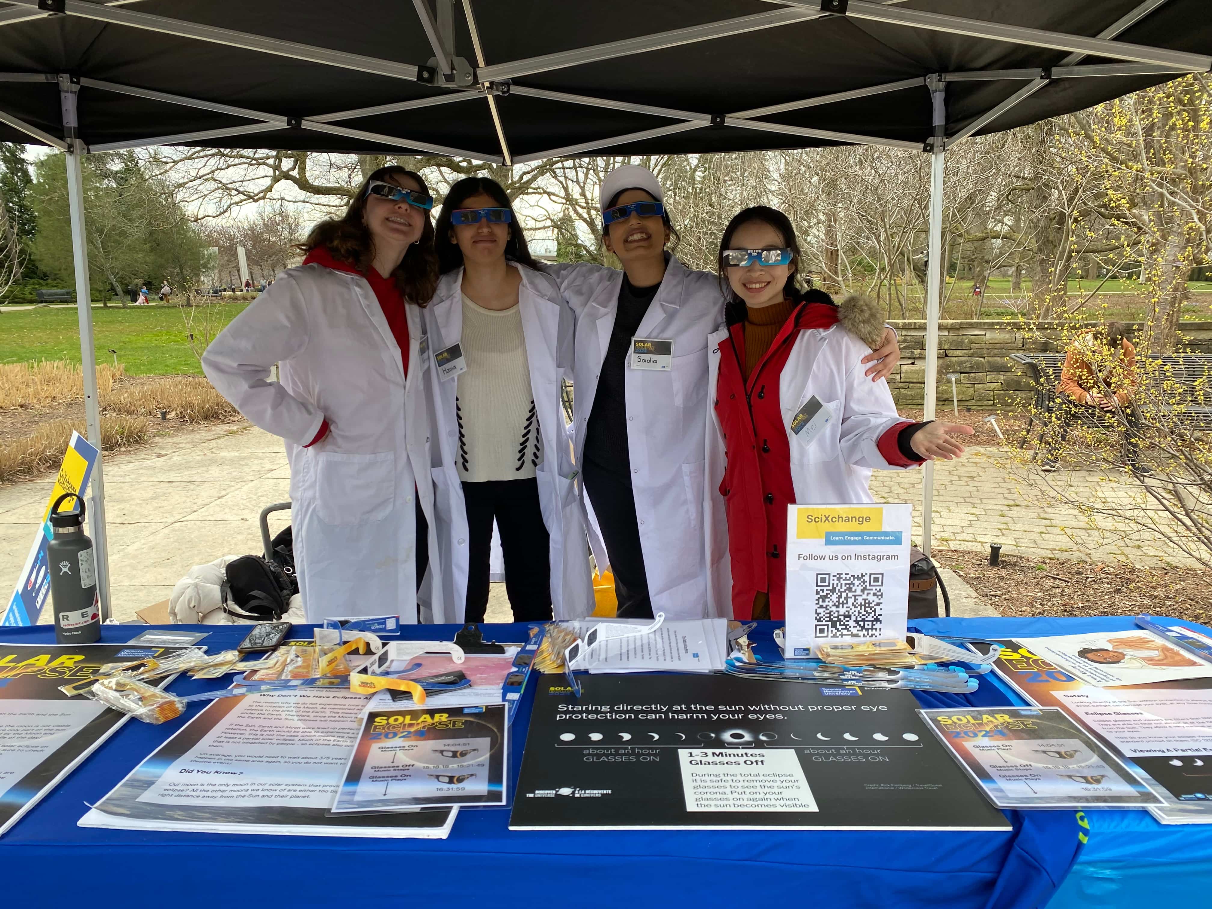 Four SciXchange volunteers in white lab coats pose for a photo under the eclipse booth at the Butterfly Conservatory in Niagara Falls.